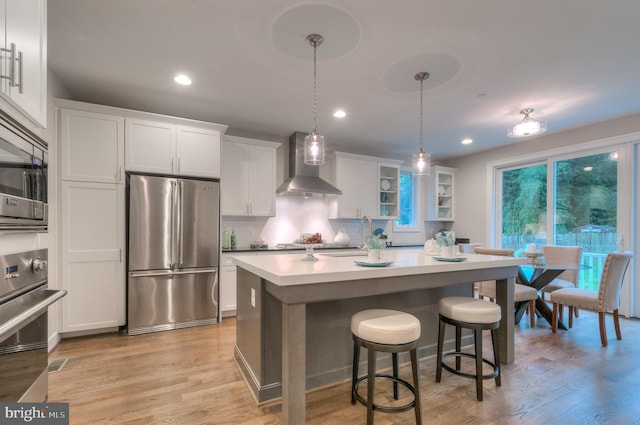 kitchen with wall chimney exhaust hood, hanging light fixtures, stainless steel appliances, white cabinetry, and light hardwood / wood-style floors