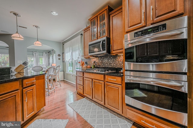 kitchen with tasteful backsplash, appliances with stainless steel finishes, hanging light fixtures, dark stone countertops, and light hardwood / wood-style flooring