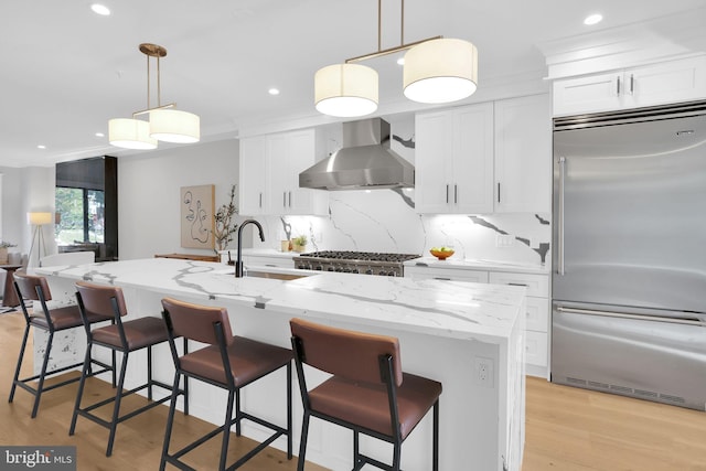 kitchen featuring wall chimney range hood, recessed lighting, white cabinets, light stone countertops, and stainless steel built in fridge