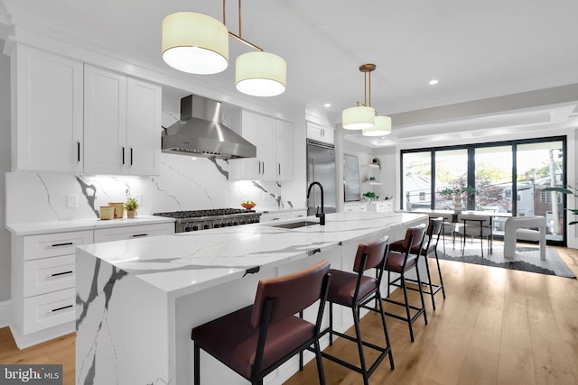kitchen with built in fridge, a sink, white cabinetry, wall chimney range hood, and a center island with sink