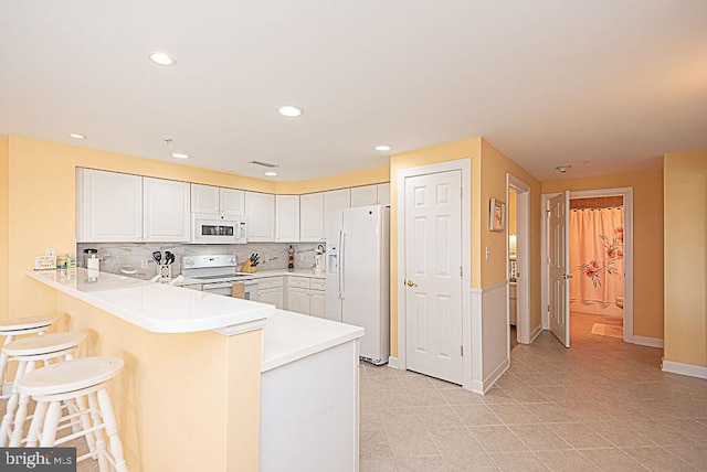 kitchen with a breakfast bar area, kitchen peninsula, white cabinetry, white appliances, and tasteful backsplash