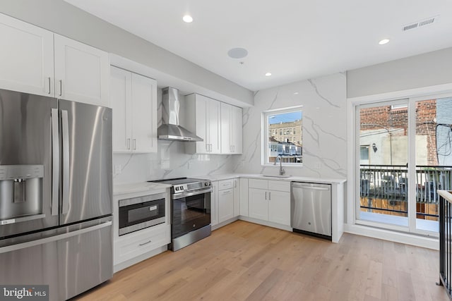 kitchen with light stone countertops, wall chimney range hood, light wood-type flooring, stainless steel appliances, and white cabinets