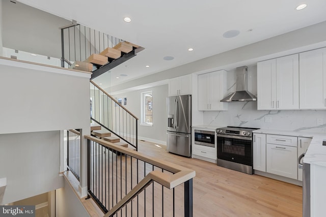 kitchen with wall chimney range hood, decorative backsplash, appliances with stainless steel finishes, white cabinetry, and light hardwood / wood-style floors