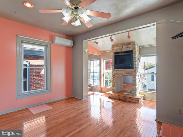 unfurnished living room with rail lighting, ceiling fan, light hardwood / wood-style flooring, an AC wall unit, and a stone fireplace