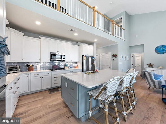 kitchen featuring a high ceiling, an island with sink, stainless steel appliances, white cabinetry, and light hardwood / wood-style floors