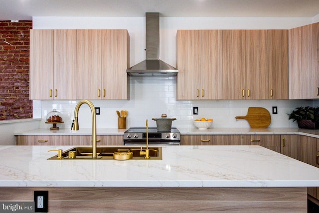 kitchen featuring wall chimney exhaust hood, stainless steel range, brick wall, light brown cabinetry, and light stone counters