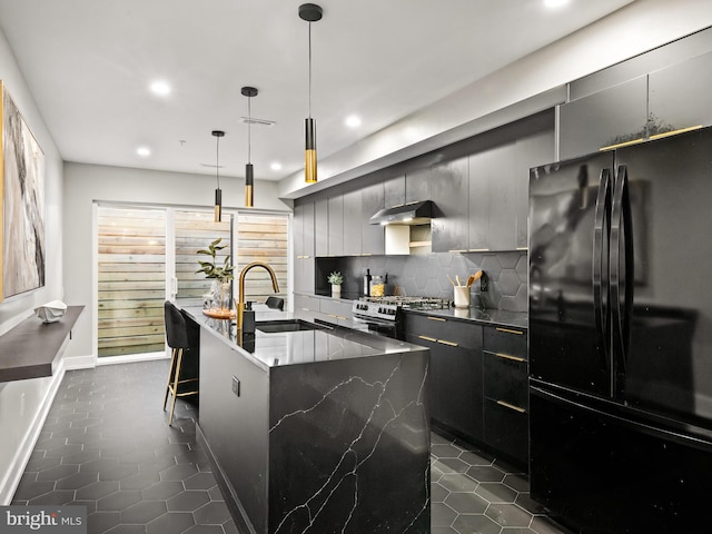 kitchen featuring gas range, black fridge, an island with sink, sink, and decorative light fixtures