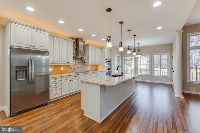 kitchen featuring light stone countertops, appliances with stainless steel finishes, wall chimney exhaust hood, pendant lighting, and a kitchen island with sink