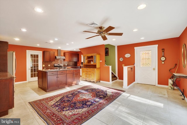 kitchen with ceiling fan, backsplash, an island with sink, light tile patterned flooring, and wall chimney exhaust hood