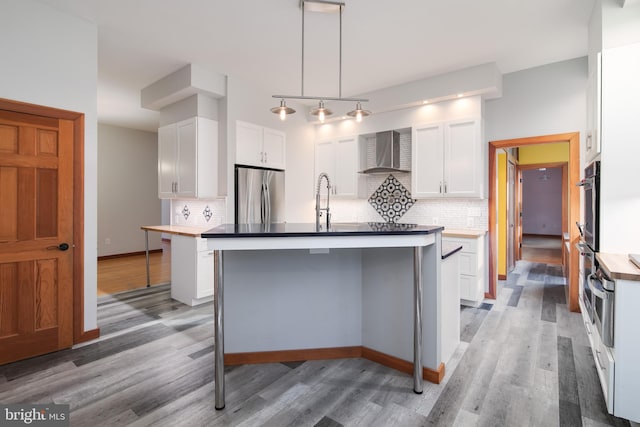 kitchen with a kitchen island with sink, white cabinets, wall chimney exhaust hood, stainless steel fridge, and tasteful backsplash