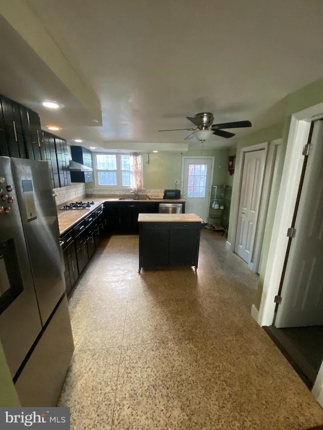 kitchen featuring wall chimney range hood, tasteful backsplash, ceiling fan, appliances with stainless steel finishes, and a kitchen island