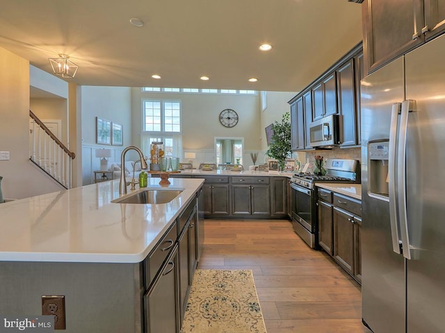 kitchen featuring a kitchen island with sink, a towering ceiling, stainless steel appliances, sink, and light hardwood / wood-style flooring