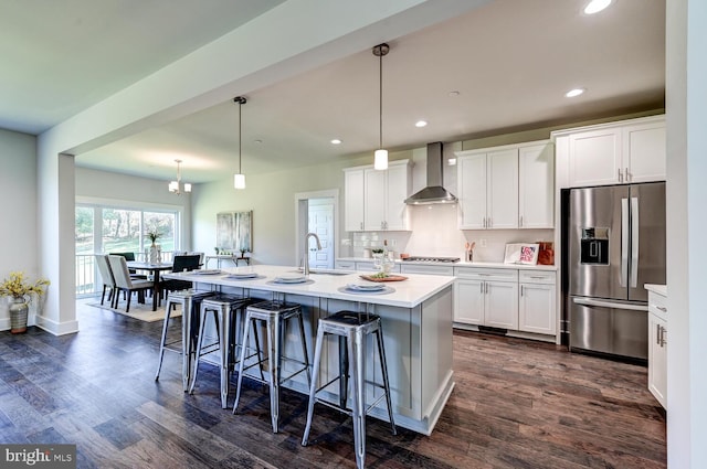 kitchen featuring wall chimney range hood, stainless steel refrigerator with ice dispenser, white cabinets, and an island with sink