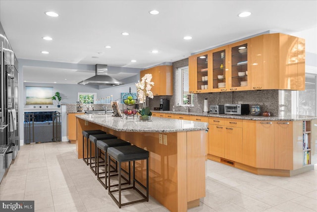 kitchen featuring light stone counters, wall chimney range hood, light tile patterned floors, and a kitchen island with sink