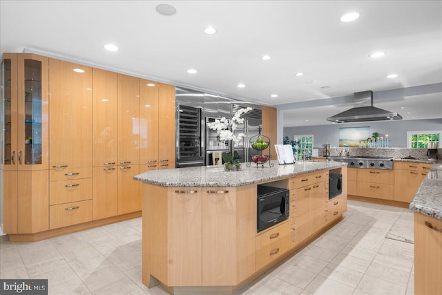 kitchen featuring light stone countertops, light brown cabinetry, a center island, wall chimney exhaust hood, and black microwave