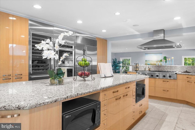 kitchen featuring light brown cabinets, wall chimney range hood, stainless steel gas cooktop, black microwave, and light stone countertops