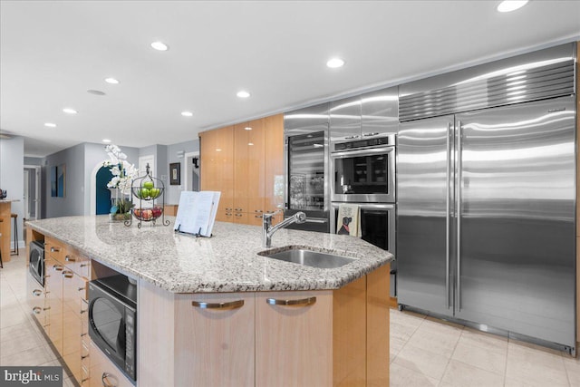 kitchen with built in appliances, light stone counters, sink, and a kitchen island with sink