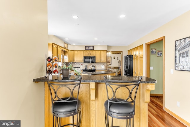 kitchen with light hardwood / wood-style flooring, kitchen peninsula, backsplash, black appliances, and a breakfast bar