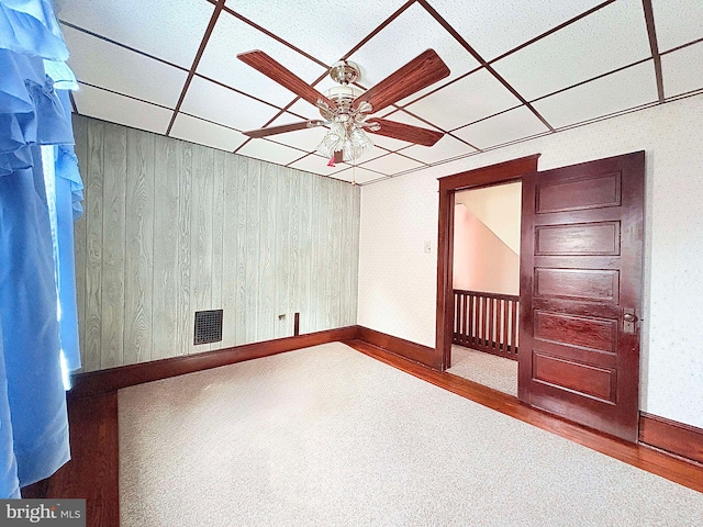 spare room featuring carpet flooring, ceiling fan, wood walls, and a drop ceiling