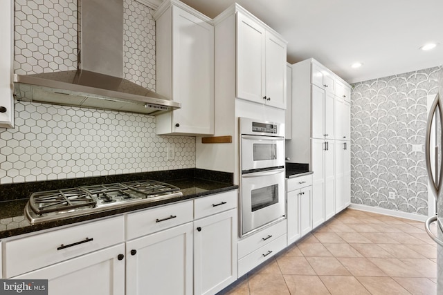 kitchen with wall chimney exhaust hood, stainless steel appliances, light tile patterned floors, dark stone countertops, and white cabinets