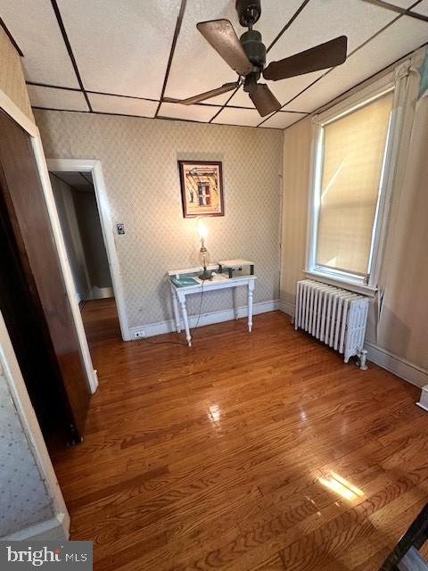 empty room featuring a paneled ceiling, wood-type flooring, radiator heating unit, and ceiling fan