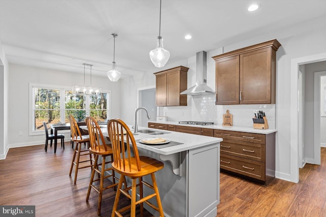 kitchen featuring sink, dark wood-type flooring, hanging light fixtures, wall chimney range hood, and a center island with sink