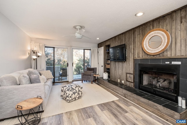 living room with light wood-type flooring, a textured ceiling, a tiled fireplace, ceiling fan, and wooden walls