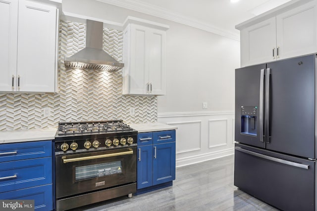 kitchen featuring appliances with stainless steel finishes, blue cabinetry, crown molding, white cabinets, and wall chimney range hood