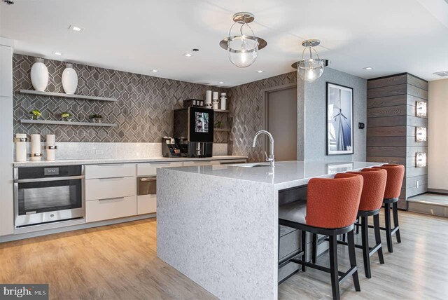 kitchen featuring stainless steel oven, hanging light fixtures, a center island with sink, light wood-type flooring, and white cabinetry