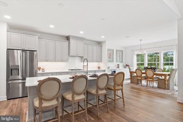 kitchen featuring an inviting chandelier, light wood-type flooring, a center island with sink, and stainless steel fridge