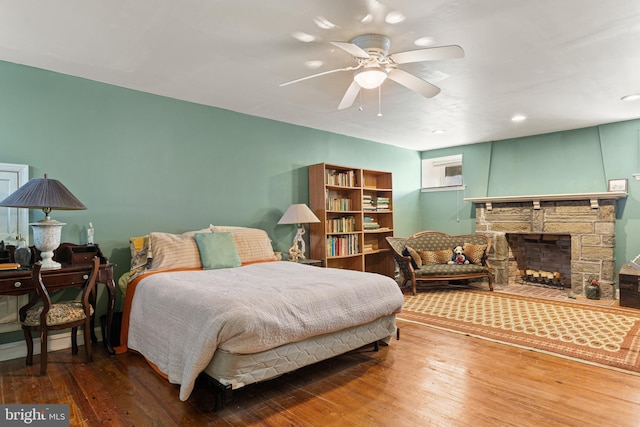 bedroom with a stone fireplace, wood-type flooring, and ceiling fan