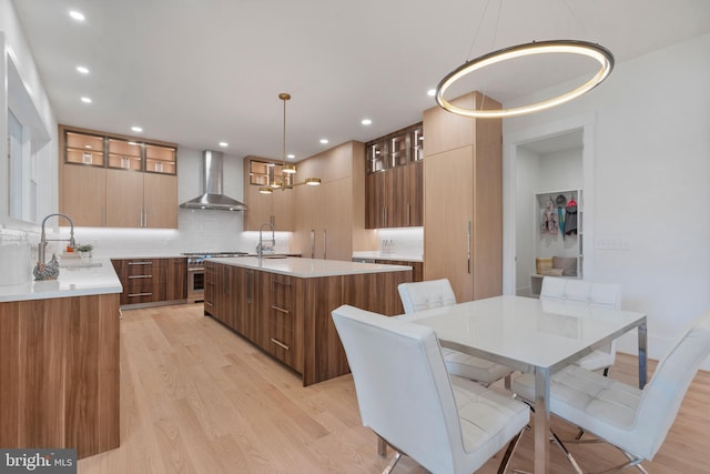 kitchen with a large island with sink, wall chimney exhaust hood, light hardwood / wood-style flooring, hanging light fixtures, and sink