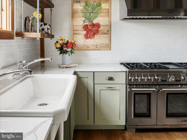 kitchen with wall chimney range hood, green cabinetry, light stone countertops, hardwood / wood-style floors, and stainless steel range