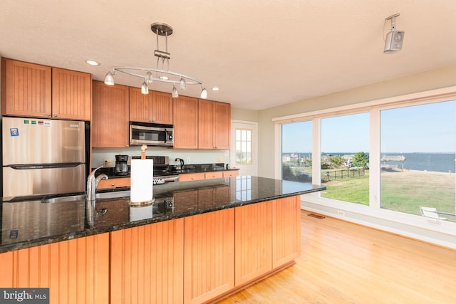 kitchen featuring appliances with stainless steel finishes, light hardwood / wood-style floors, dark stone counters, pendant lighting, and a water view