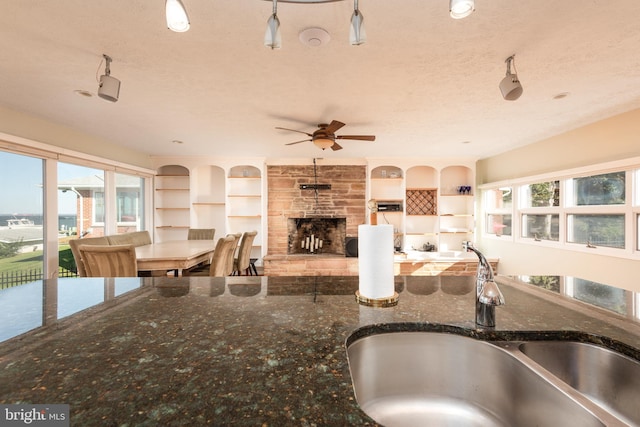 kitchen with ceiling fan, a textured ceiling, dark stone counters, a stone fireplace, and sink