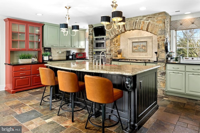 kitchen with stone tile floors, hanging light fixtures, glass insert cabinets, a kitchen island with sink, and a chandelier