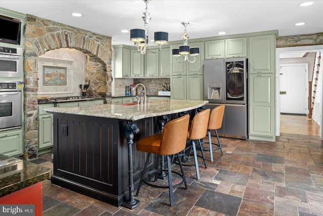 kitchen with hanging light fixtures, green cabinets, appliances with stainless steel finishes, an island with sink, and light stone countertops