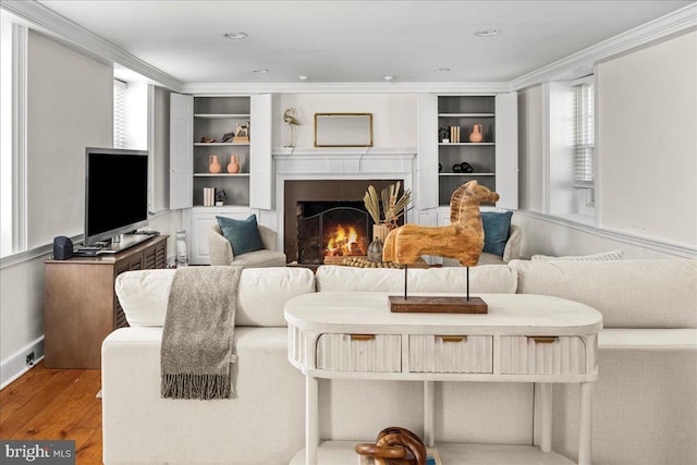 living room featuring built in shelves, recessed lighting, ornamental molding, light wood-type flooring, and a warm lit fireplace