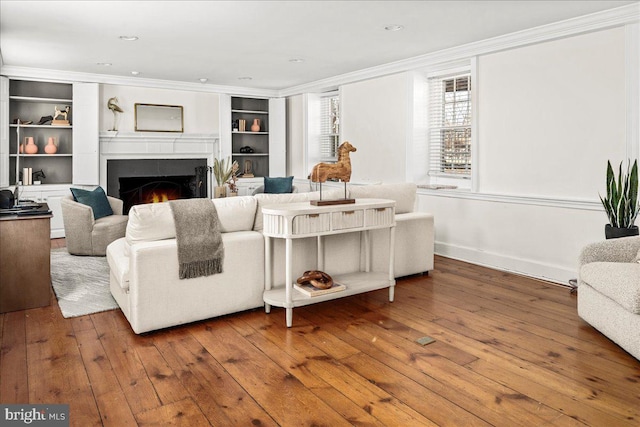 living area featuring baseboards, wood finished floors, a lit fireplace, crown molding, and built in shelves