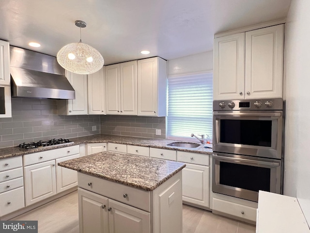 kitchen featuring wall chimney range hood, hanging light fixtures, sink, appliances with stainless steel finishes, and white cabinetry