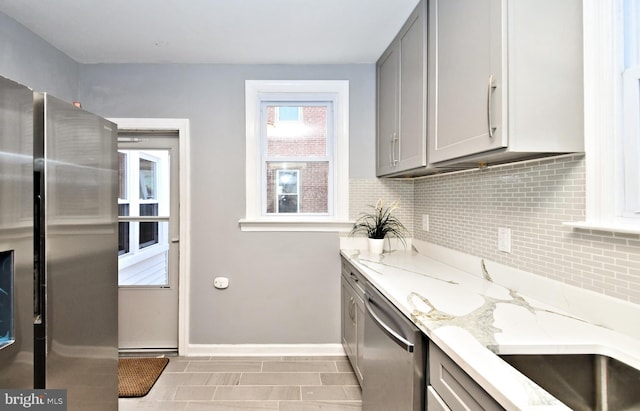 kitchen with stainless steel appliances, gray cabinetry, light stone counters, decorative backsplash, and baseboards