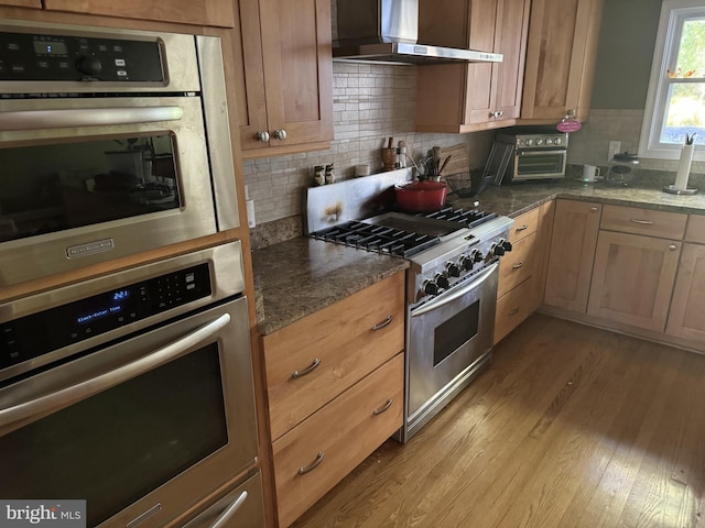 kitchen with appliances with stainless steel finishes, dark stone countertops, ventilation hood, and light wood-type flooring