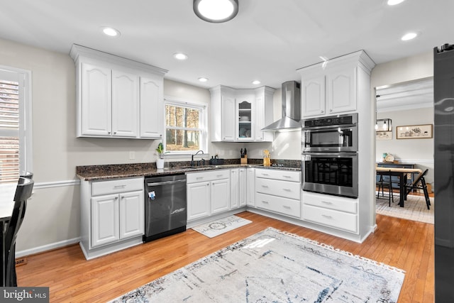 kitchen with wall chimney exhaust hood, stainless steel appliances, light hardwood / wood-style flooring, dark stone countertops, and white cabinets