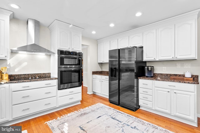 kitchen with white cabinetry, wall chimney exhaust hood, dark stone counters, black appliances, and light wood-type flooring