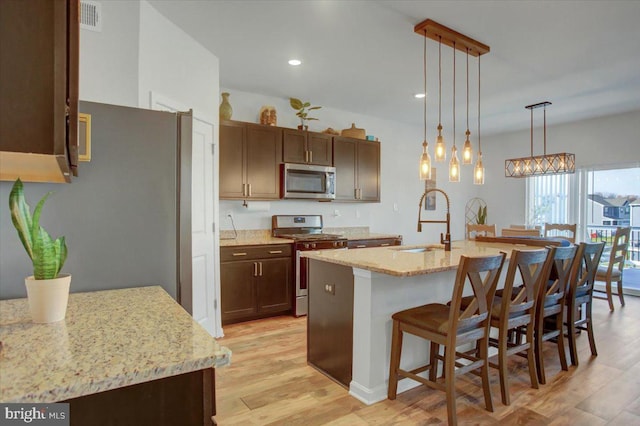 kitchen featuring a kitchen island with sink, sink, light wood-type flooring, appliances with stainless steel finishes, and decorative light fixtures