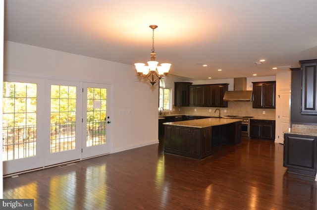 kitchen featuring a kitchen island with sink, wall chimney exhaust hood, dark brown cabinets, pendant lighting, and dark hardwood / wood-style flooring