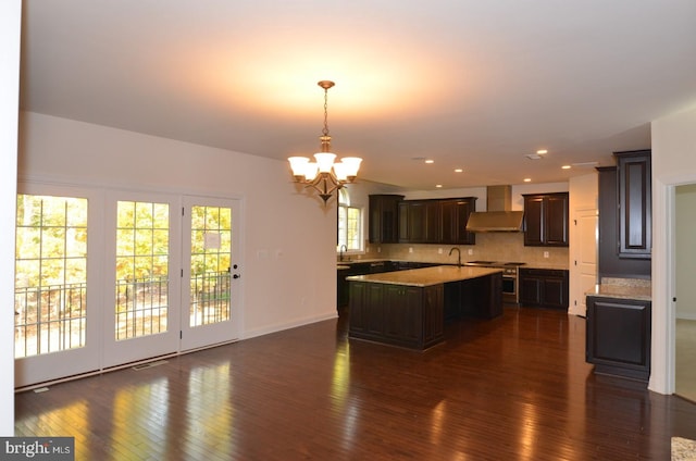 kitchen featuring a center island with sink, hanging light fixtures, dark wood-type flooring, wall chimney exhaust hood, and dark brown cabinetry