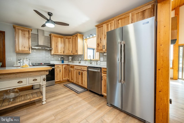 kitchen featuring wall chimney range hood, backsplash, light wood-type flooring, appliances with stainless steel finishes, and ceiling fan