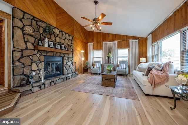 living room with ceiling fan, high vaulted ceiling, light hardwood / wood-style flooring, a stone fireplace, and wooden walls