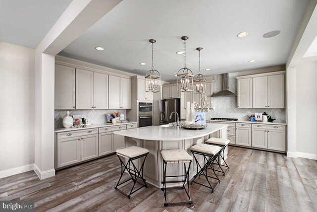 kitchen with appliances with stainless steel finishes, hardwood / wood-style floors, wall chimney exhaust hood, a breakfast bar, and a kitchen island with sink